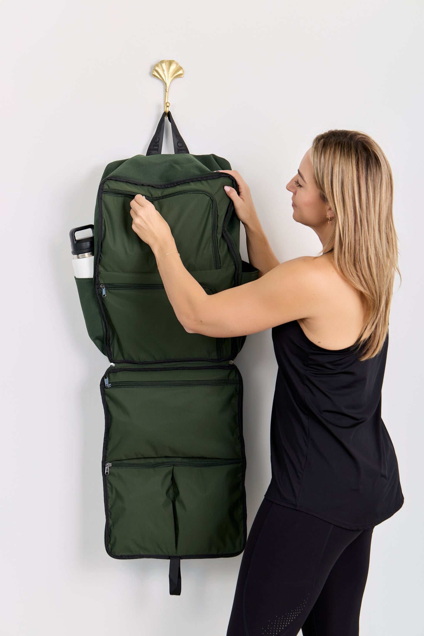 Person hanging a green backpack on a hook against a white wall